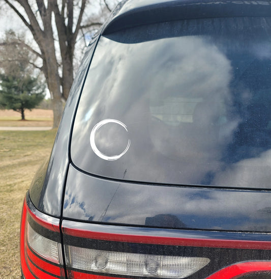Car window with a Enso Circle decal and reflection of trees and sky.
